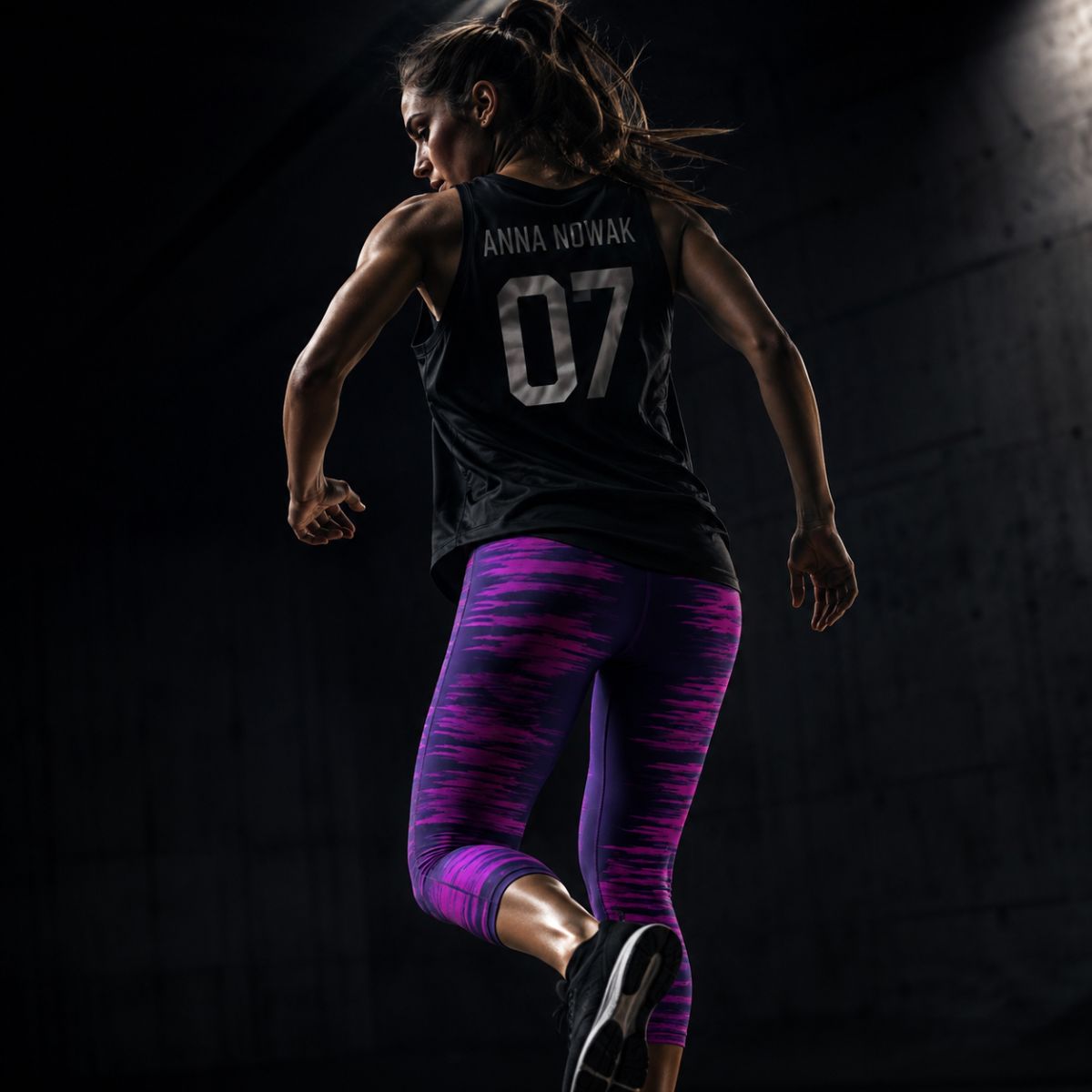 Female athlete in mid-air, back turned, wearing a black tank with 'ANNA NOWAK' and '07' and purple-pink leggings in a dark setting